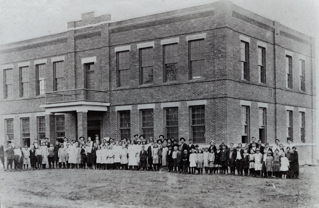 Elementary school students standing in front of a two-story brick schoolhouse, photo in black and white.